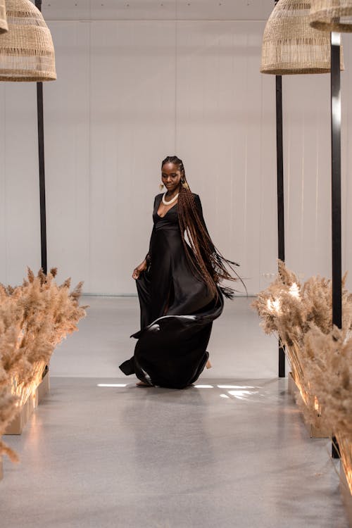 a black woman with long braids is posing in a flowing black maxi dress in a stylish studio setting. the background includes modern lamps and pampas grass decor, creating an elegant and sophisticated atmosphere, reminiscent of a fashion runway or photoshoot. the model's pose is graceful, and the dress's movement is captured beautifully.