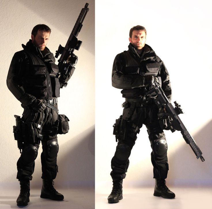 this image shows a man dressed as a soldier in black tactical gear standing in a studio holding a large gun in each hand. he is looking directly at the camera.  the background is a light color.