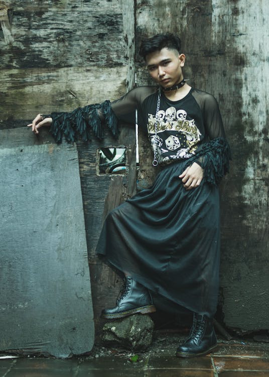 young person with short dark hair is posing against a weathered wooden wall. they are wearing a black sheer longsleeved top with a skull and dice graphic, a long black skirt, and black doc martens. a long black necklace with skull charms hangs around their neck. the overall style is grunge, alternative, and gothinspired, with a dark and edgy aesthetic. the photo has a moody, atmospheric quality.