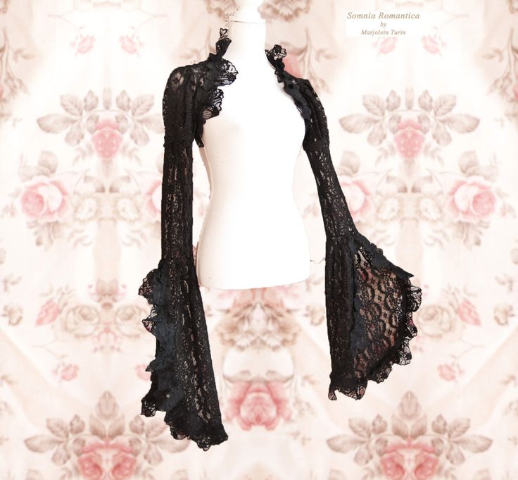 a black lace bolero with bell sleeves is displayed on a white mannequin against a floral patterned background. the bolero has a delicate lace pattern and ruffled edges around the neck and sleeves. the background features a vintagestyle floral design with roses. the label "somnia romantica by marjolein turin" is visible in the upper right corner.