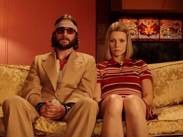 a man and woman sit on a couch together. the man is wearing a brown suit with a brown headband and sunglasses. he is looking to the right. the woman is wearing a red and white striped dress. she is looking straight ahead with a serious expression. they are in what appears to be a living room, with a retro 1970s aesthetic. the background is out of focus.