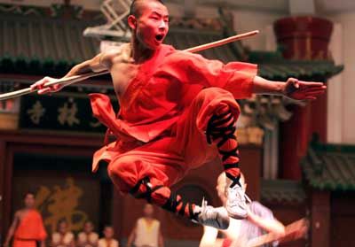 a young shaolin monk in red training gear is midair, midkick. he is in the middle of performing a martial arts technique. the staff he is holding is at his side, and he is focused on the target ahead.  the photograph was taken in a training facility.