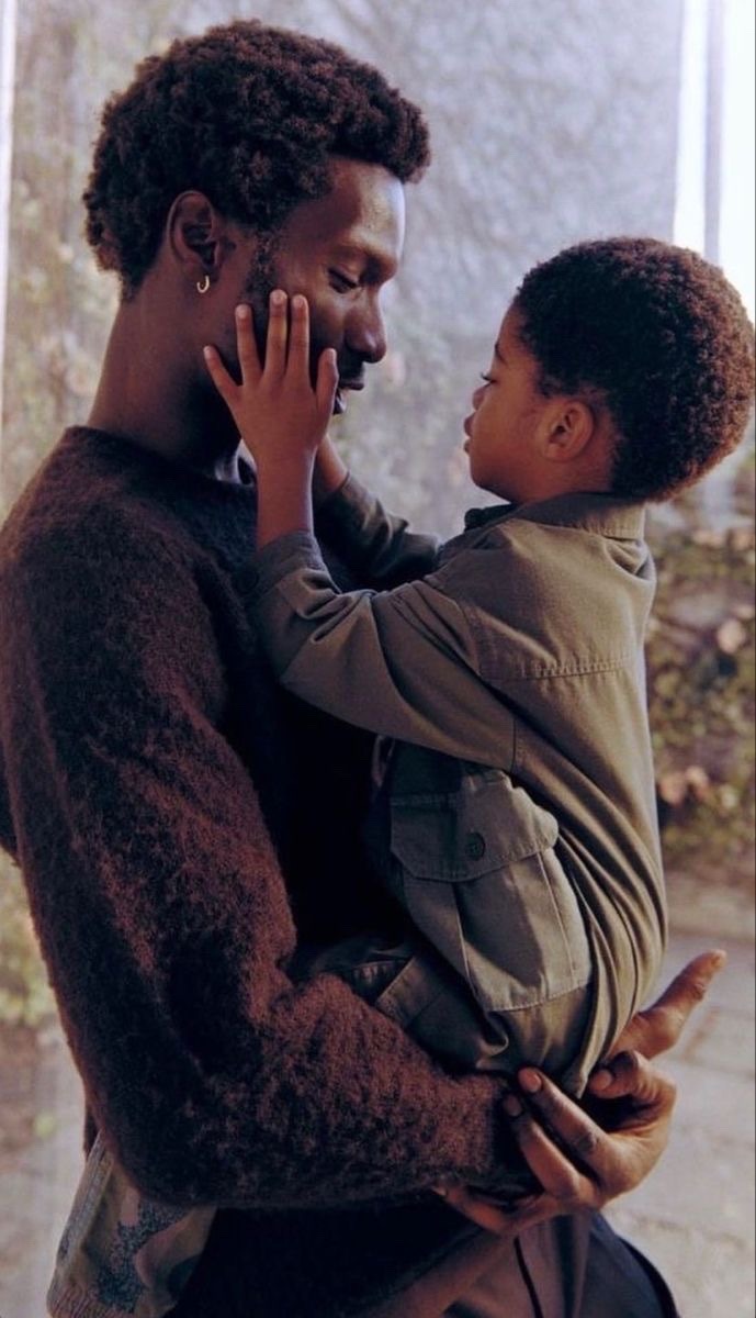 a black man and his young son are embracing in a loving hug. the father is wearing a brown sweater and the son is wearing a green jacket. the father is looking down at his son with a soft and loving expression. the image is a beautiful depiction of the love and bond between a father and son.