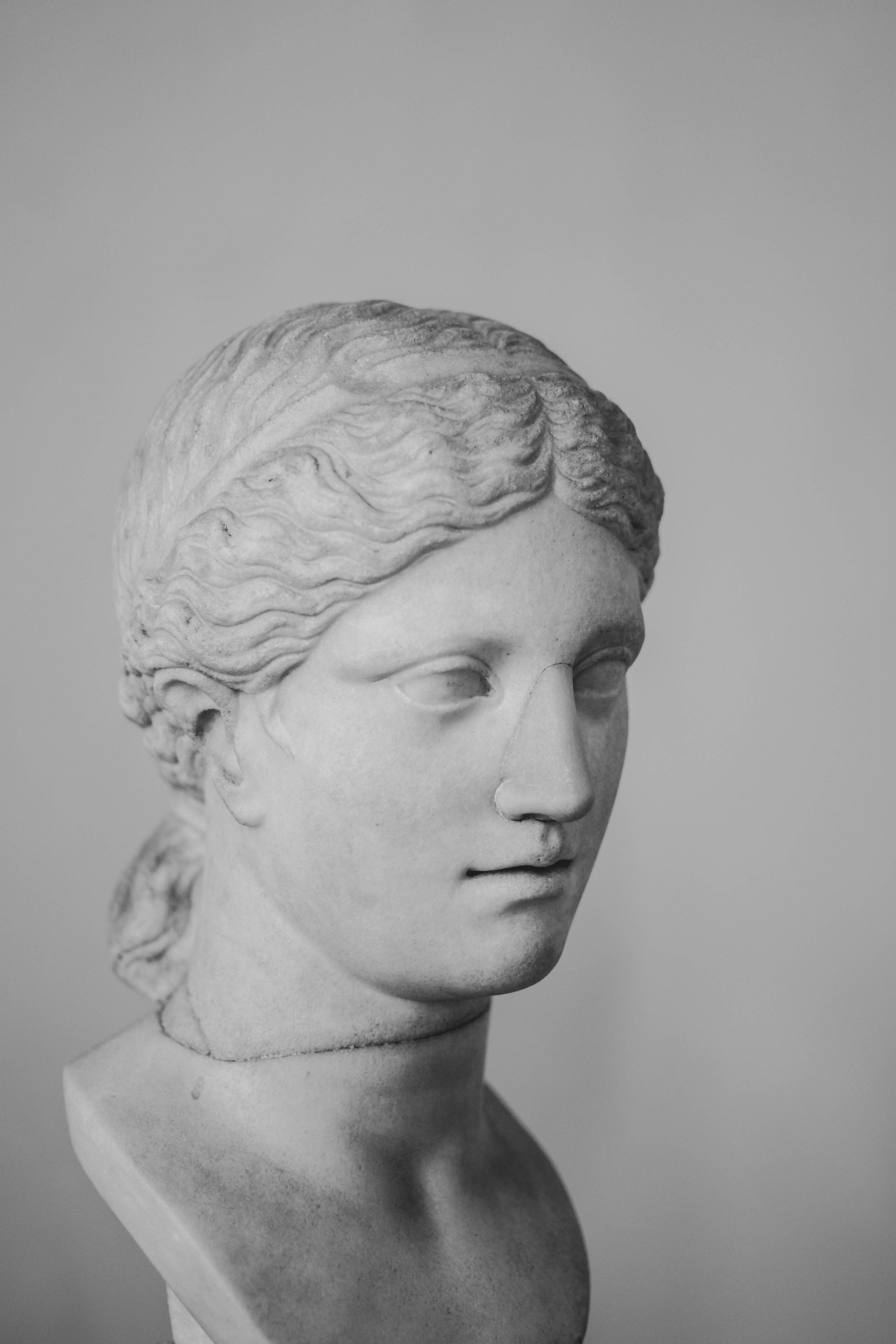 a black and white image of a marble bust of a woman. the bust is of a classical style, possibly greek or roman, and depicts a woman with her hair pulled back in a bun. the sculpture is lit softly and evenly, highlighting the smooth curves of the marble. the background is a plain white wall, which further emphasizes the subject.