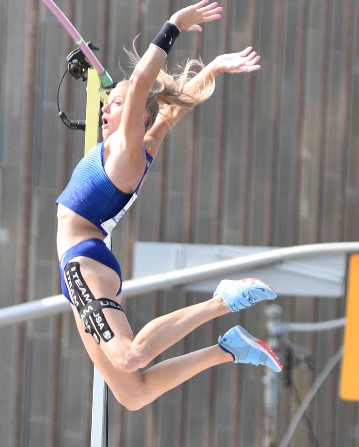 a female pole vaulter in midair during a competition, she has a determined look on her face as she reaches for the bar.  she is wearing a blue and white uniform and has black tape on her leg, showing her dedication and strength. she has blonde hair and is wearing blue and white shoes, making her a vibrant presence in the scene.  the background is out of focus and features a grey concrete wall and a yellow sign.