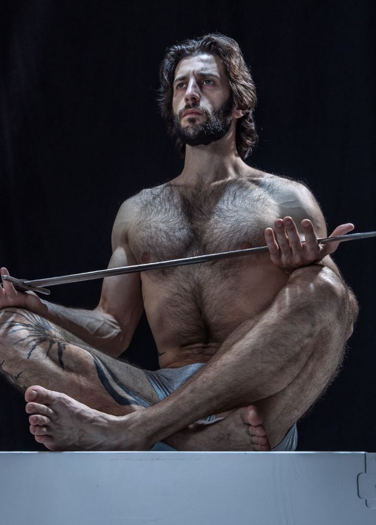 a muscular man with chest hair is sitting down with his legs crossed. he is looking up at the camera with a serious expression and is holding a sharp metal sword in his hands. his body is bare from the waist up. the background is dark and shadowy, adding to the dramatic feel of the image.