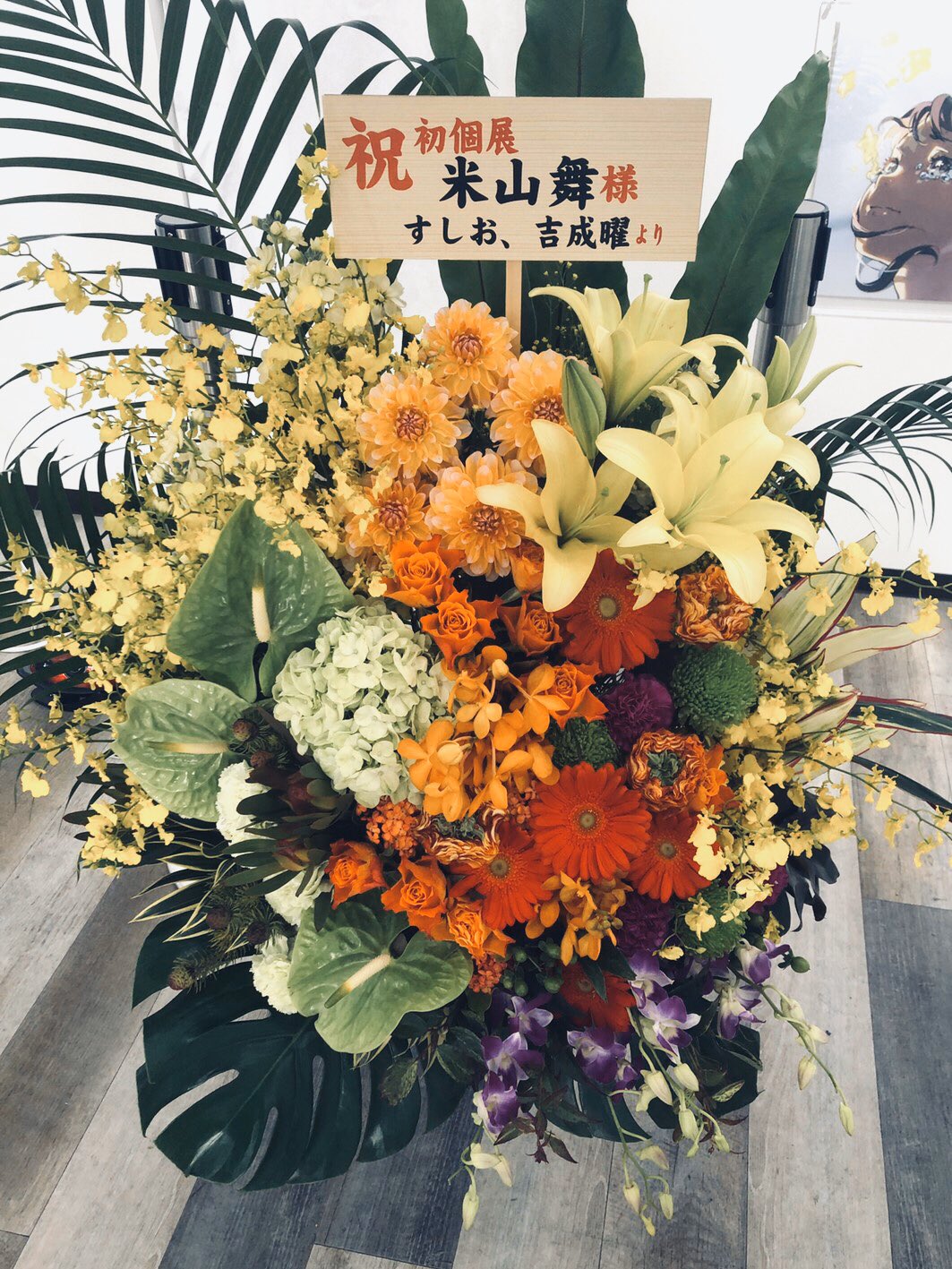 a vibrant floral arrangement stands on a light colored floor in front of a white wall. the arrangement features yellow, orange, and red flowers, including lilies, orchids, and gerbera daisies. a wooden sign with japanese text congratulating someone on the opening of a new business is displayed prominently among the blooms. the scene is brightly lit, likely with natural light coming from the left side of the frame.