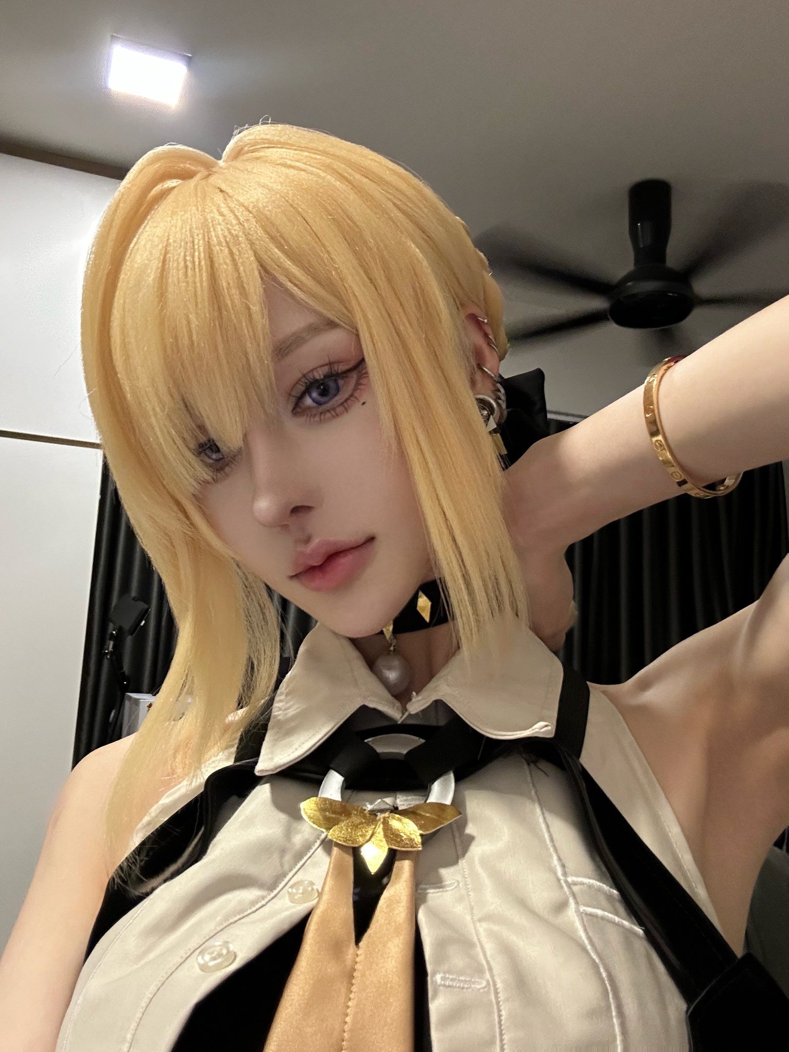 closeup selfie of a young woman cosplaying a character with long blonde hair styled in bangs. she's wearing a beige shirt and vestlike outfit with gold and black accessories, including a unique gold tielike piece.  she has on several pieces of jewelry: earrings, necklace, and a bracelet. the background shows a portion of a room with a ceiling fan. the overall style is anime or mangainspired. the photo appears to be a selfportrait taken indoors.