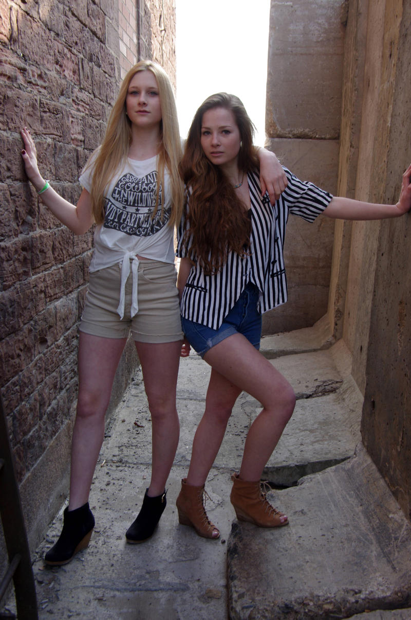 the image shows two teenage girls standing in a narrow alleyway between a brick wall and a concrete wall.  one girl has blonde hair and is wearing a beige pair of shorts and a white tshirt with text. the other girl has brown hair and wears a black and white striped shirt and denim shorts. both girls are wearing ankle boots, one pair brown and the other black. they are posing together, arms around each other,  suggesting friendship.  the alleyway is lit by natural sunlight. the overall feel is casual and summery.