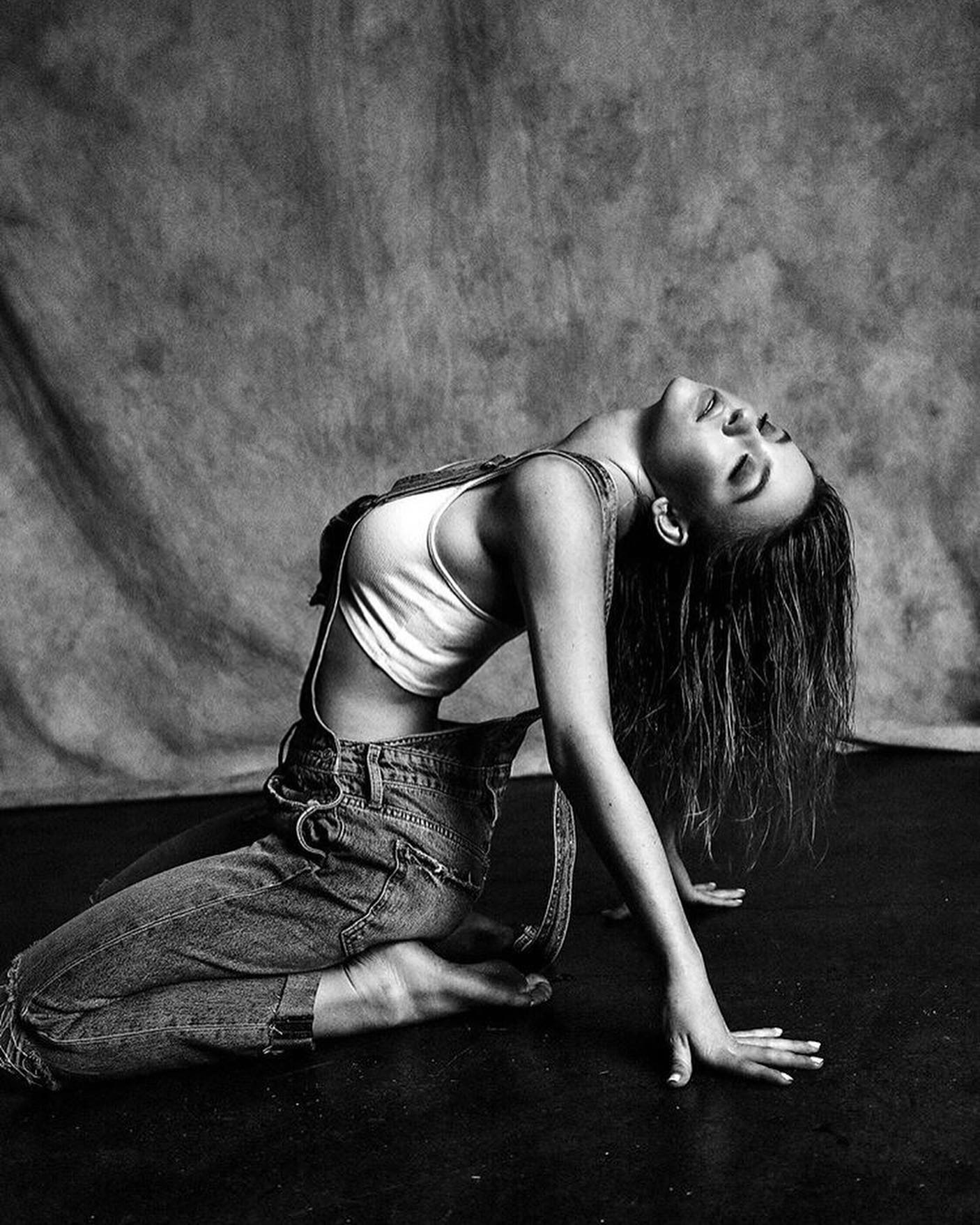 the black and white photograph showcases a young woman in a sensual pose. she is positioned on the floor, kneeling with her legs slightly apart, her body arched backward, and her head tilted upwards.  she wears a white cropped top and loosefitting denim overalls. her hair is long and falls around her shoulders. the background is a dark, textured backdrop that contrasts with her lighter skin and clothing. the overall mood is artistic and dramatic, enhanced by the monochrome palette and strategic lighting. the image emphasizes her body shape and posture.