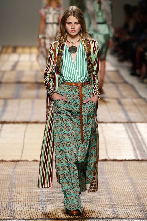a fashion model walks the runway wearing a stylish bohemian outfit. the outfit consists of a long striped coat in red, green, and brown over a teal paisley print palazzo pants and a mint green striped top.  the model is accessorized with a large circular pendant necklace and a brown leather belt. the background shows other models and a textured runway. the overall style is chic and elegant with a bohemian flair. the image is welllit and professionally photographed.