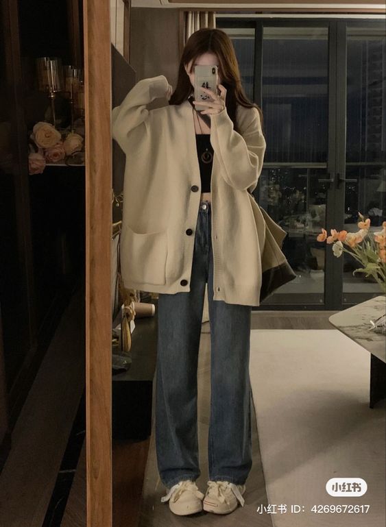 a young woman wearing a beige cardigan and blue jeans is standing in a room. she's looking into a mirror and holding her phone up to take a picture of herself. the room is decorated in a cozy and casual style. she is wearing white shoes to complete her look.