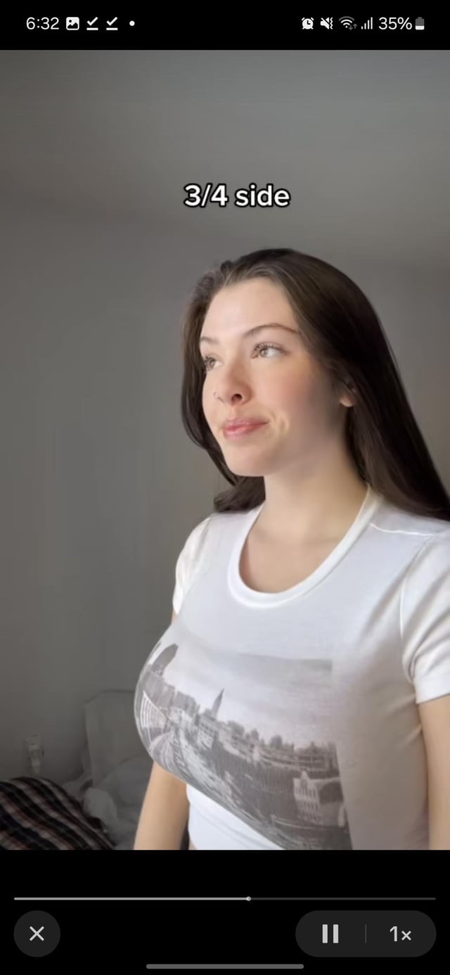 the image is a threequarter view side profile portrait of a young woman with long brown hair. she is wearing a white tshirt with a grayscale city skyline graphic printed on the front. she appears to be indoors against a neutral gray backdrop, looking slightly away from the camera.  the overall mood is casual and relaxed.  the photo appears to be a still from a video, as indicated by video player controls visible at the bottom of the image.
