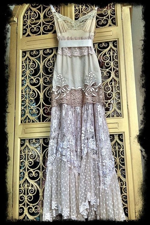 a fulllength photograph shows a beautiful, bohemianstyle wedding dress hanging on a gold ornate door. the dress is primarily beige and ivory, with layers of lace, tulle, and embroidery. the bodice is fitted with a sweetheart neckline and features delicate lace details.  a satin sash cinches the waist. the skirt is layered and flows to the floor. the background features a detailed, goldcolored door with intricate metalwork. the overall style evokes a vintage or antique aesthetic with a romantic and shabbychic feel.