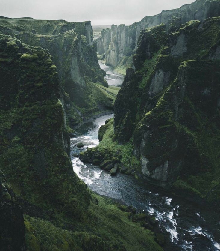 a scenic aerial shot of a canyon with a river flowing through it. the canyon walls are covered in lush green moss and the river flows through the rugged landscape. the sky is overcast.