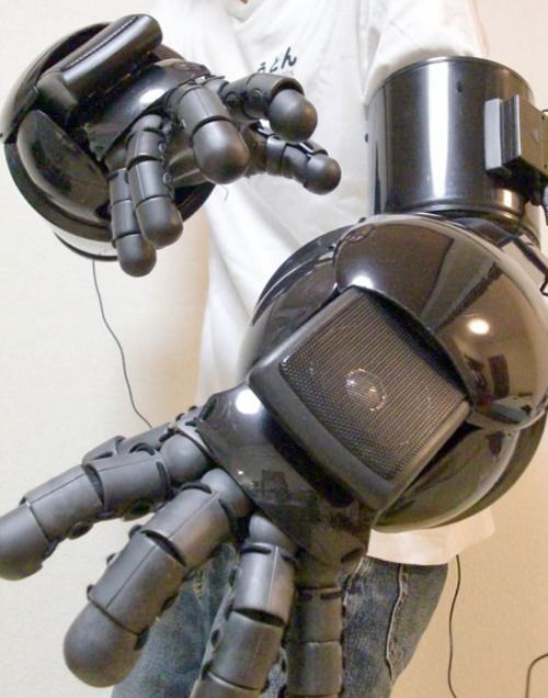 a person is wearing a futuristic, black, robotic hand on their right arm. the hand has multiple articulated fingers, and a large, speakerlike component on the back. it appears to be a robotic prosthetic. the person is wearing a white shirt and blue jeans. the background is a plain white wall.