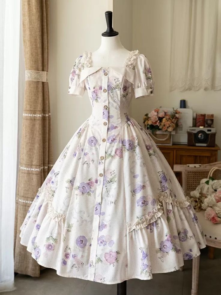 a white floral lolita dress is shown on a mannequin. the dress features puff sleeves, a square neckline, a fitted bodice with buttons down the front, and a full skirt with floral print. the dress is set against a white curtain and a wooden chair.