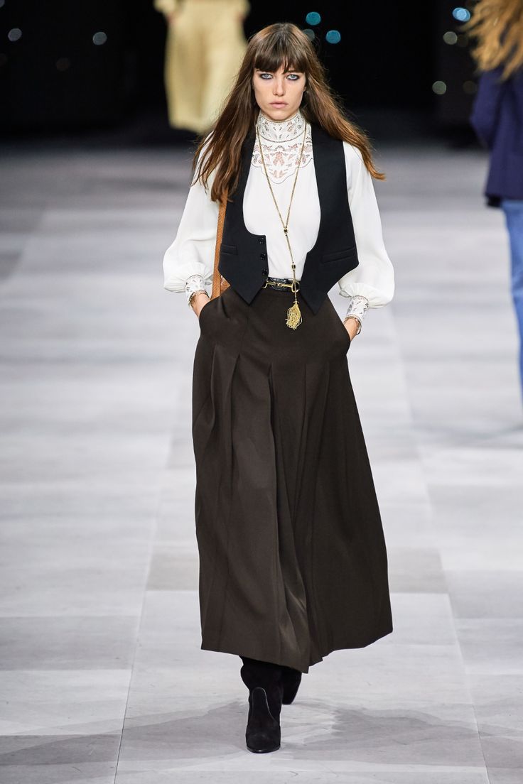 a fashion model walks down a runway in a stylish outfit. she is wearing a white blouse with a black vest and widelegged black trousers. the model is also wearing black boots, a necklace with a pendant, and earrings. her hair is styled in a straight, long, parted, dark brunette style with bangs. she has  simple makeup with a natural shade of lipstick.