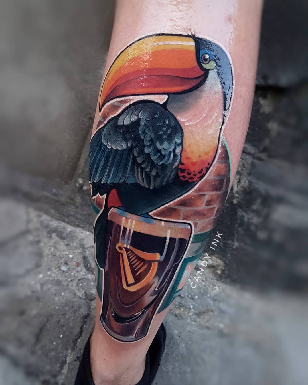a color tattoo of a toucan perched on a glass of guinness beer is displayed on someone's lower leg. the toucan's black feathers, orange beak, and white chest are brightly colored against the dark beer and muted brick background. the guinness logo is clearly visible on the glass.  the tattoo was done by an artist credited as "ciady ink."