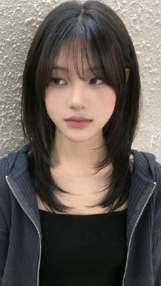 a portrait of a young asian woman with a trendy mediumlength hairstyle and wispy bangs. she is wearing a black top and a dark gray zipup hoodie. she has soft, natural makeup and a neutral expression. the lighting is soft and diffused. the image is taken from an eyelevel perspective.