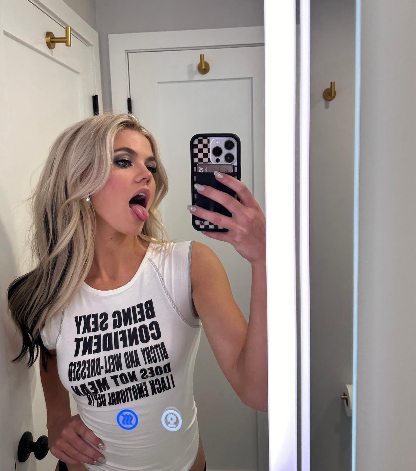 a woman is taking a selfie in a bathroom mirror, sticking her tongue out. she is wearing a white shirt with black writing on it.