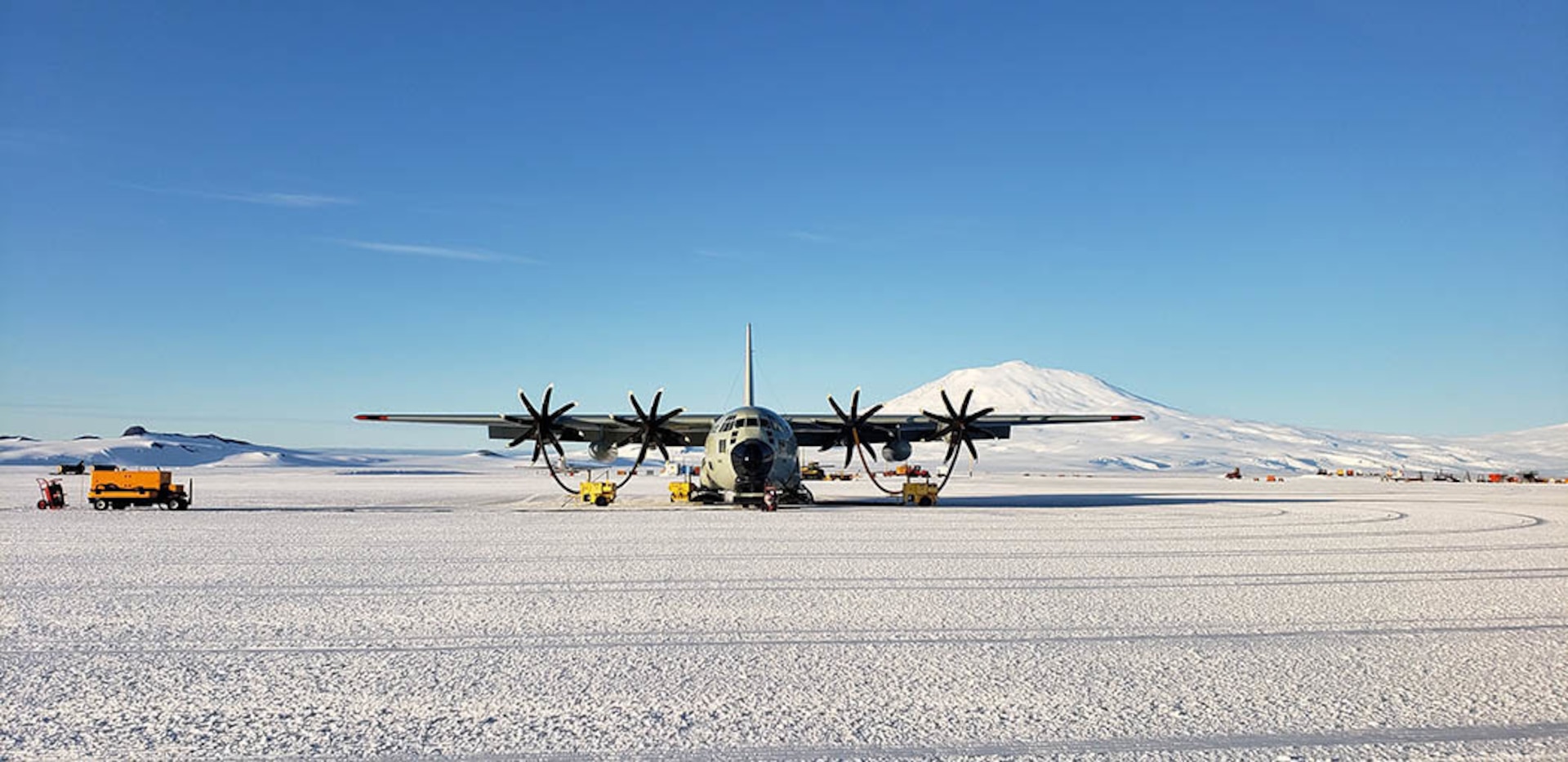 a c130 hercules cargo plane is parked on a snowy runway in antarctica. the plane is being refueled by a yellow fuel truck. in the background, there are mountains covered in snow. the sky is blue and clear.  the ground is covered in snow, and the snow is textured and very bright white. the horizon is clear, and the sky is very clear. the image is welllit and shows the plane and the surrounding area in detail.  the airplane's propellers are folded in.