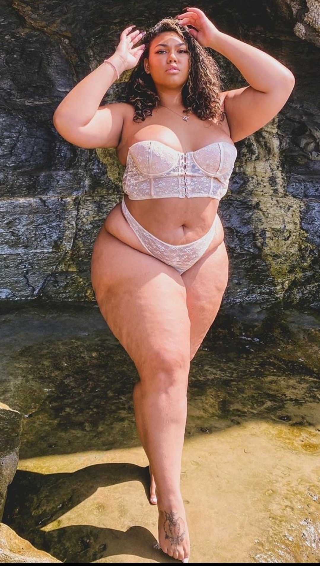 a woman in a white lace bra and panty set stands in front of a rock formation. she smiles and looks directly at the camera.