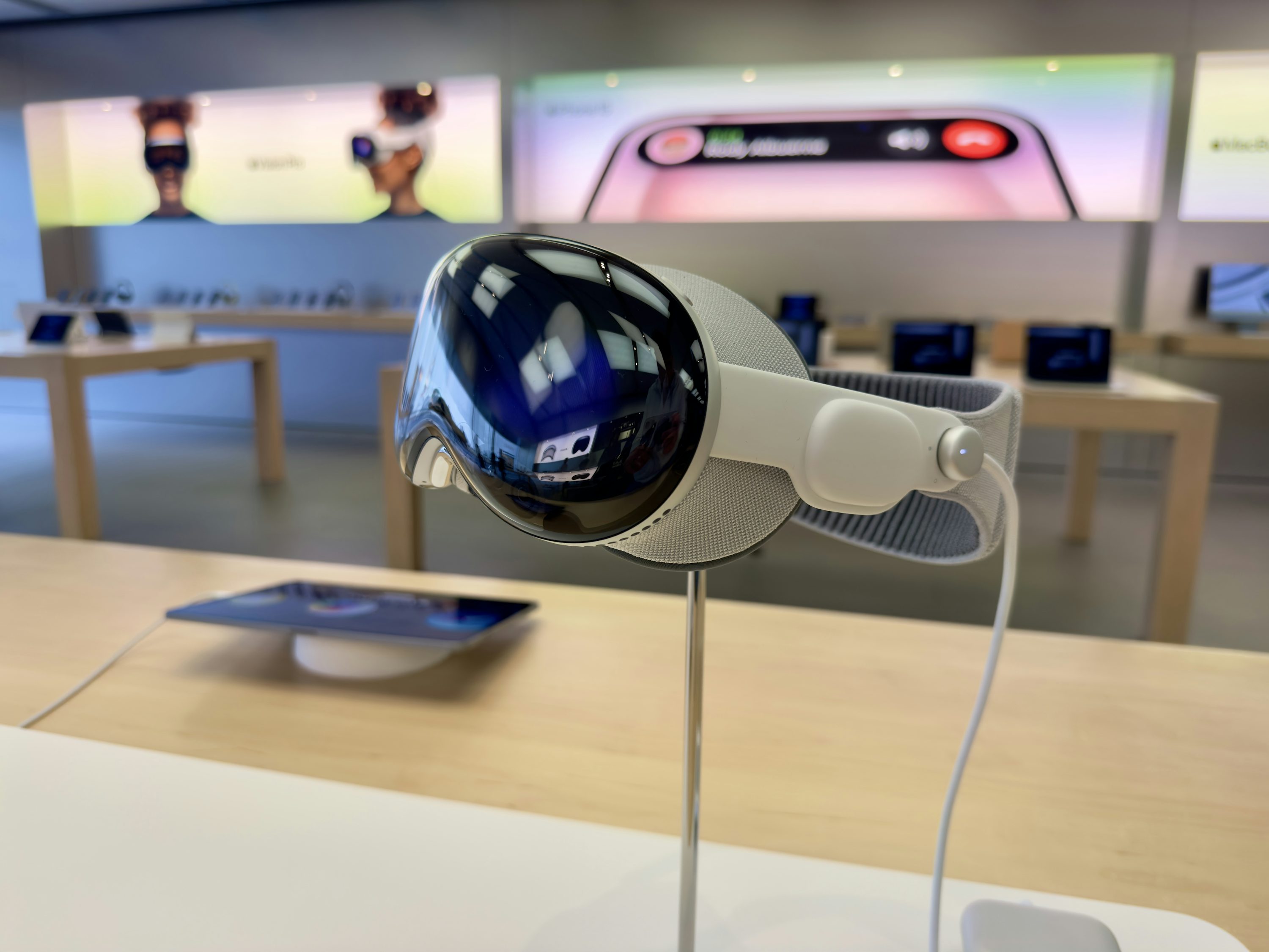 the image shows the apple vision pro vr headset on display in an apple store. the headset is positioned on a stand on a table. in the background are other tables with apple products and promotional displays. the image focuses on the design and details of the headset.