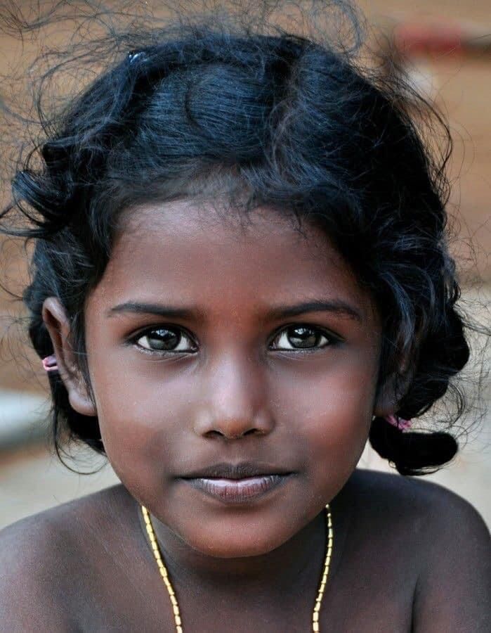 a closeup portrait of a young girl with dark skin, dark hair, and big eyes. she is wearing a gold necklace.