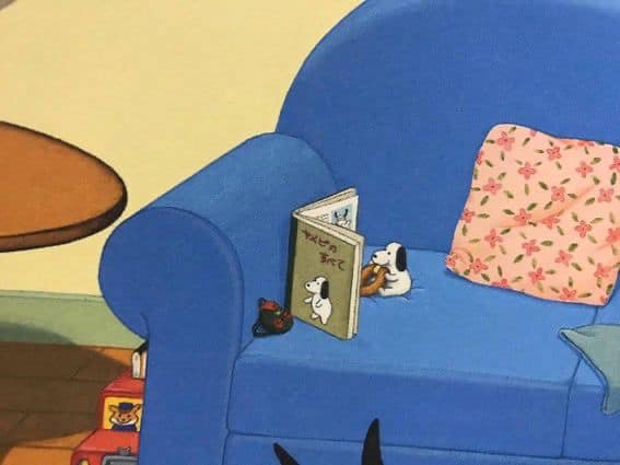 cozy illustration depicting snoopy and woodstock on a blue couch.  snoopy is holding a pretzel and sitting next to an open book written in japanese.  woodstock sits beside them. a small black object, possibly a toy or figurine, sits near the book on the couch. there's a pink floral pillow and a stack of books visible next to the couch. a wooden side table is partially visible in the background. the scene overall is very calm and inviting, suggestive of a quiet moment of reading.