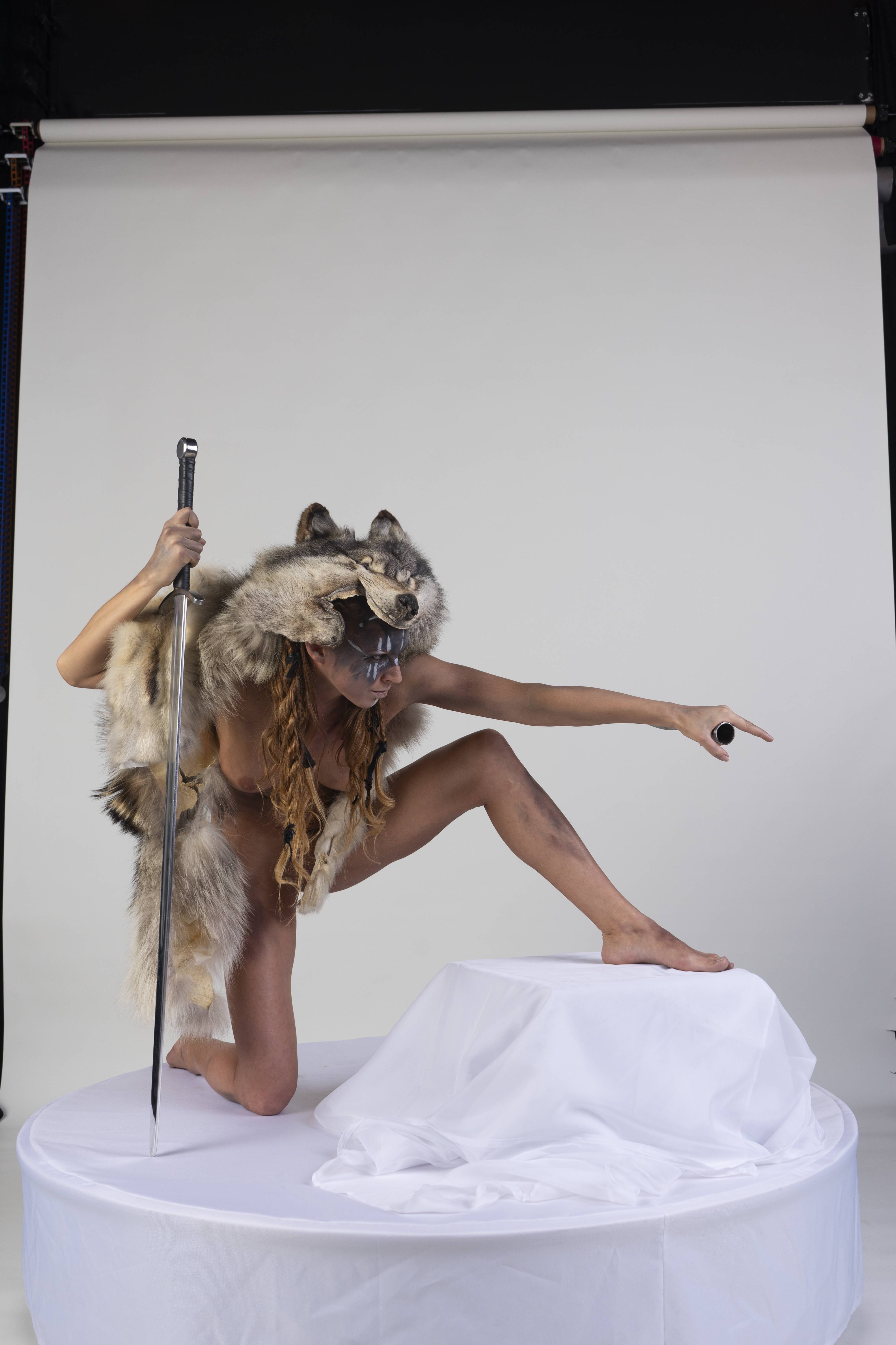 a woman in a theatrical costume poses in a studio on a white background. she is kneeling on one knee, with the other leg extended and her foot resting on a whitecovered pedestal. she wears a wolf fur headdress and loincloth, body paint, and jewelry. she holds a sword in one hand and points with the other.