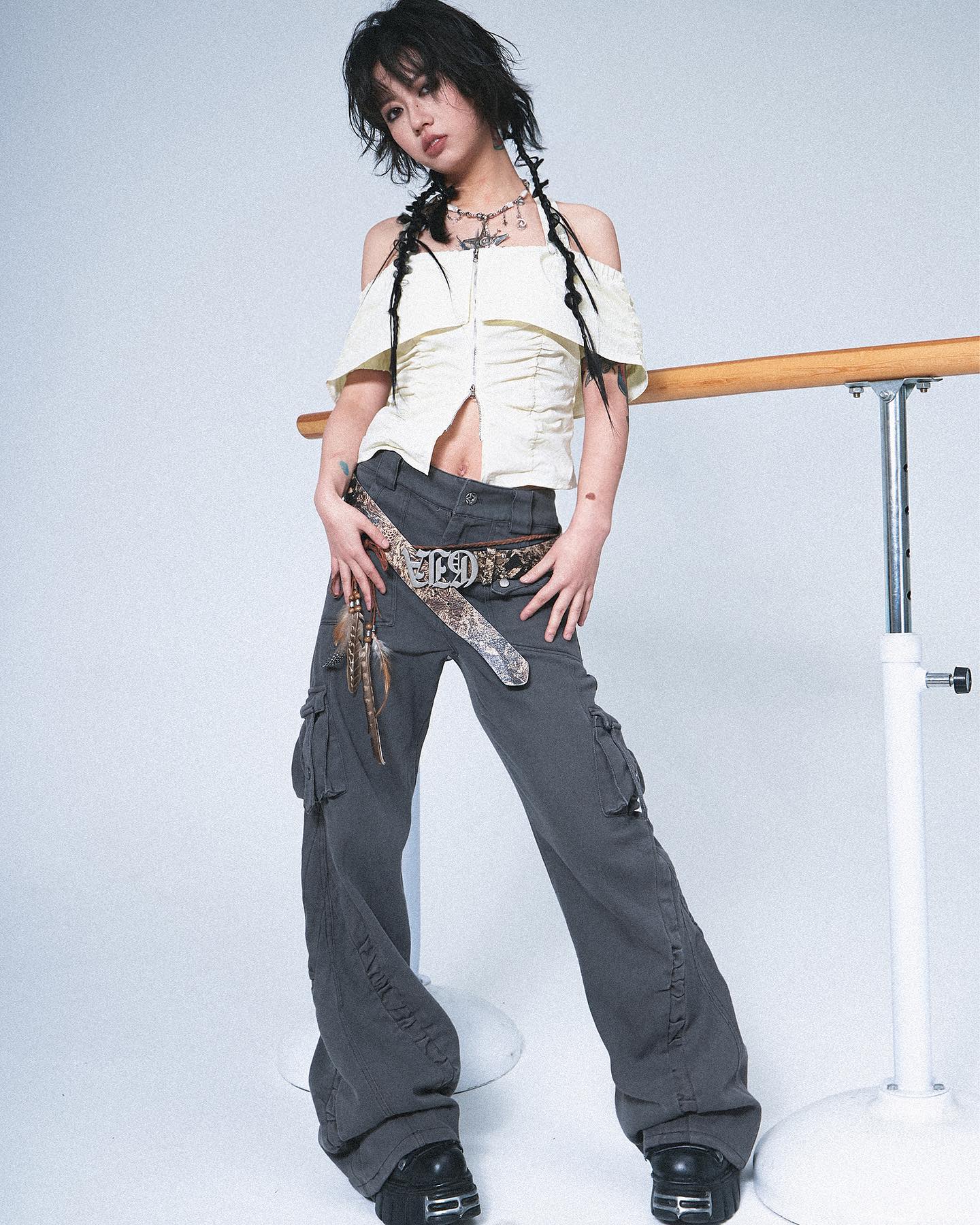a young woman with dark hair styled in braids is posing in a studio against a white background. she's wearing a pale yellow offtheshoulder top, dark grey cargo pants with a snakeskin belt, and dark boots. her overall style is edgy and alternative. a ballet barre is visible in the background.  she has a slightly defiant and confident expression.