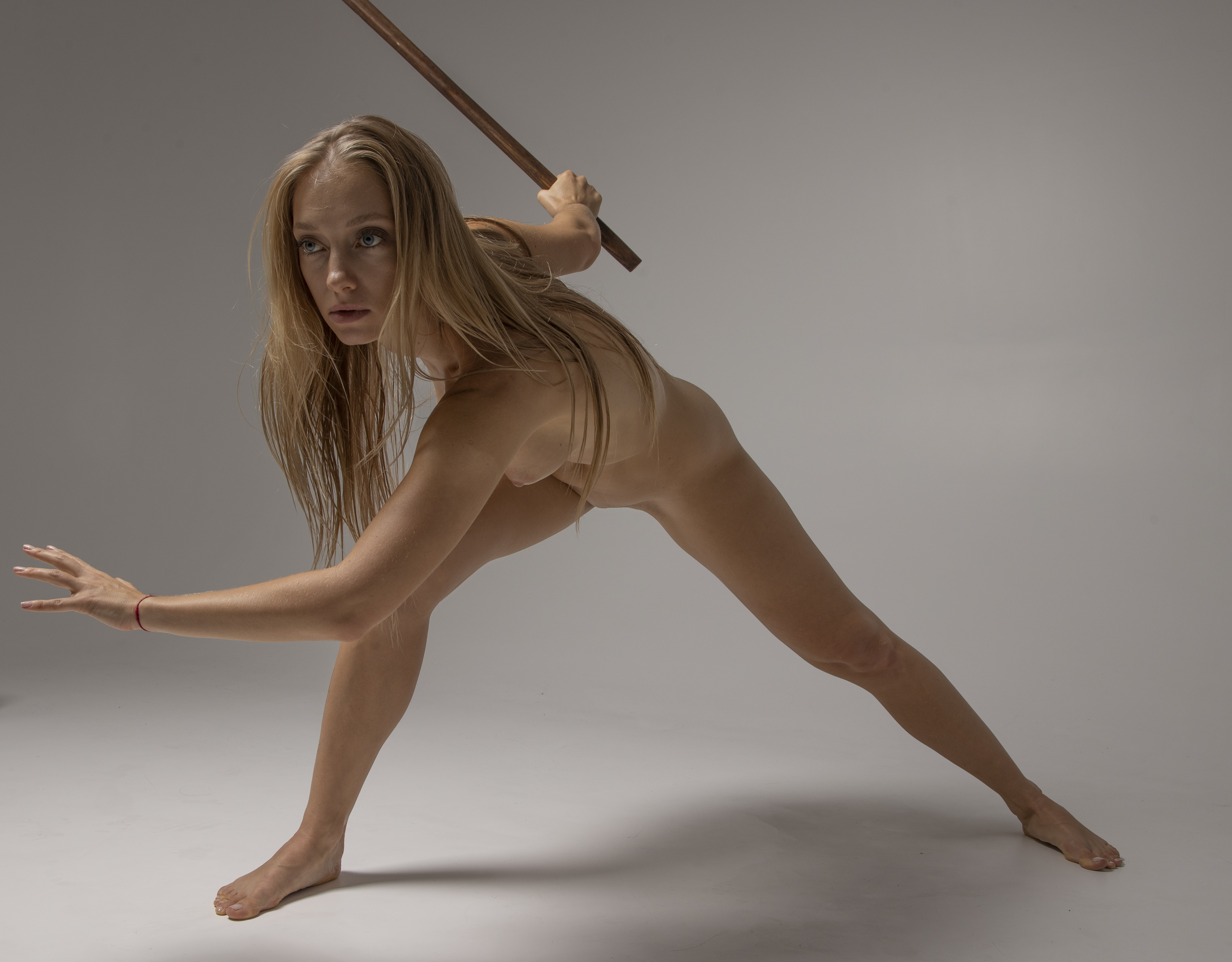 a nude woman with blonde hair is holding a wooden staff and is in a martial arts stance in a studio.
