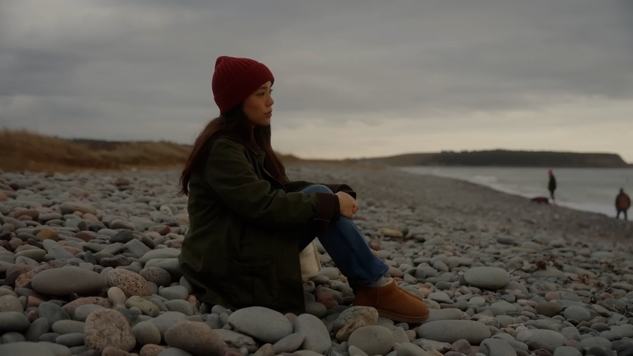 a young woman sits pensively on a rocky beach, her gaze lost in the distance. the sky is overcast, casting a soft, cool light over the scene. the colors are muted, reflecting the contemplative mood. she is bundled up in warm clothing, suggesting a chilly evening by the sea.