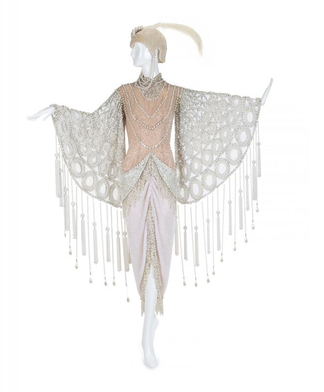 a vintage flapper dress is displayed on a mannequin. the dress is light pink and beige with a beaded and sequined overlay. the overlay has a beaded fringe. the mannequin is also wearing a beaded headpiece with a feather.
