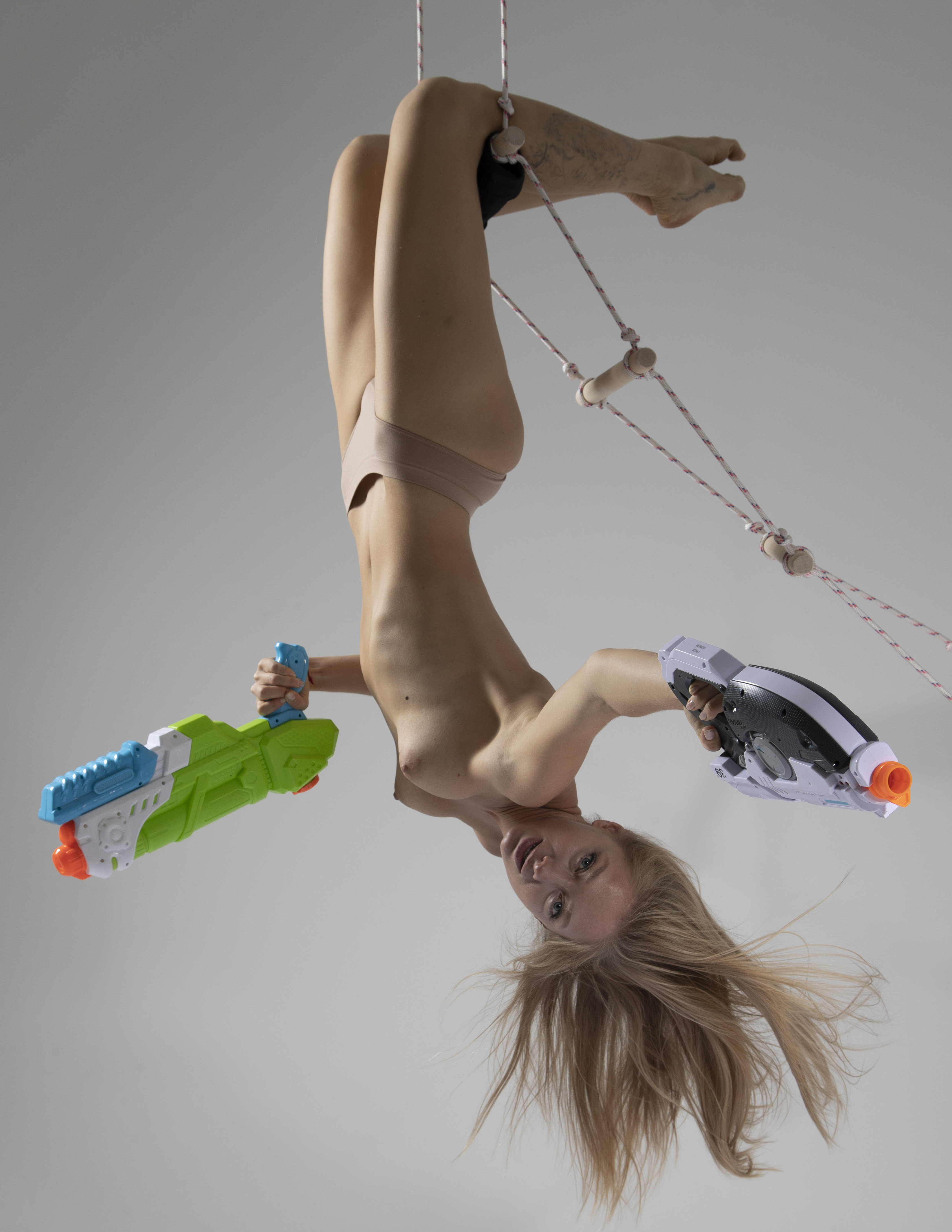 a woman is hanging upside down from a trapeze, wearing underwear, holding two squirt guns, and exposing her bare breasts. she has blonde hair that is falling downwards.