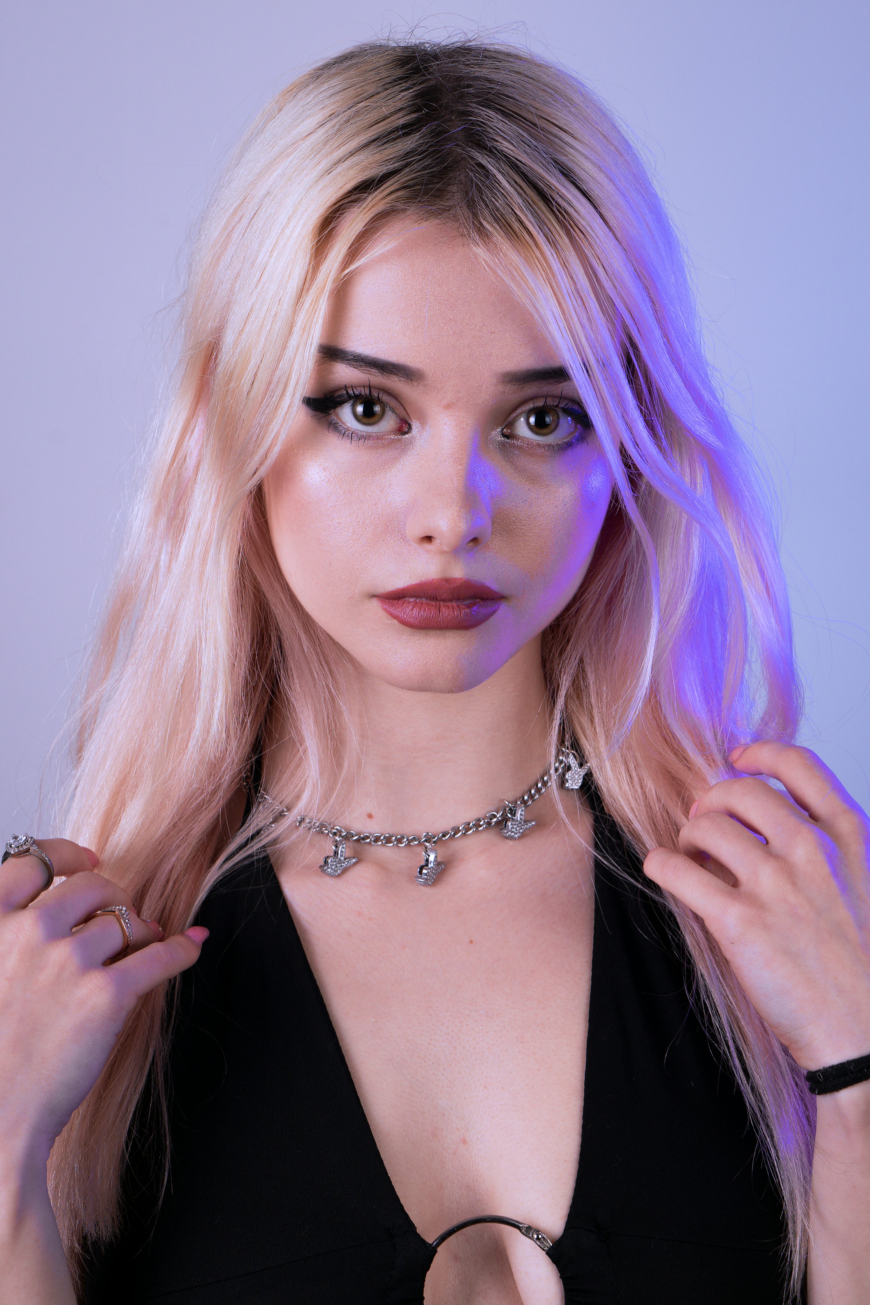 loseup portrait of a young blonde woman with striking makeup and a black top. she is wearing several silver necklaces and rings and has her hands gently touching her hair. the background is a light purple hue. the image has a fashion or beauty editorial feel.