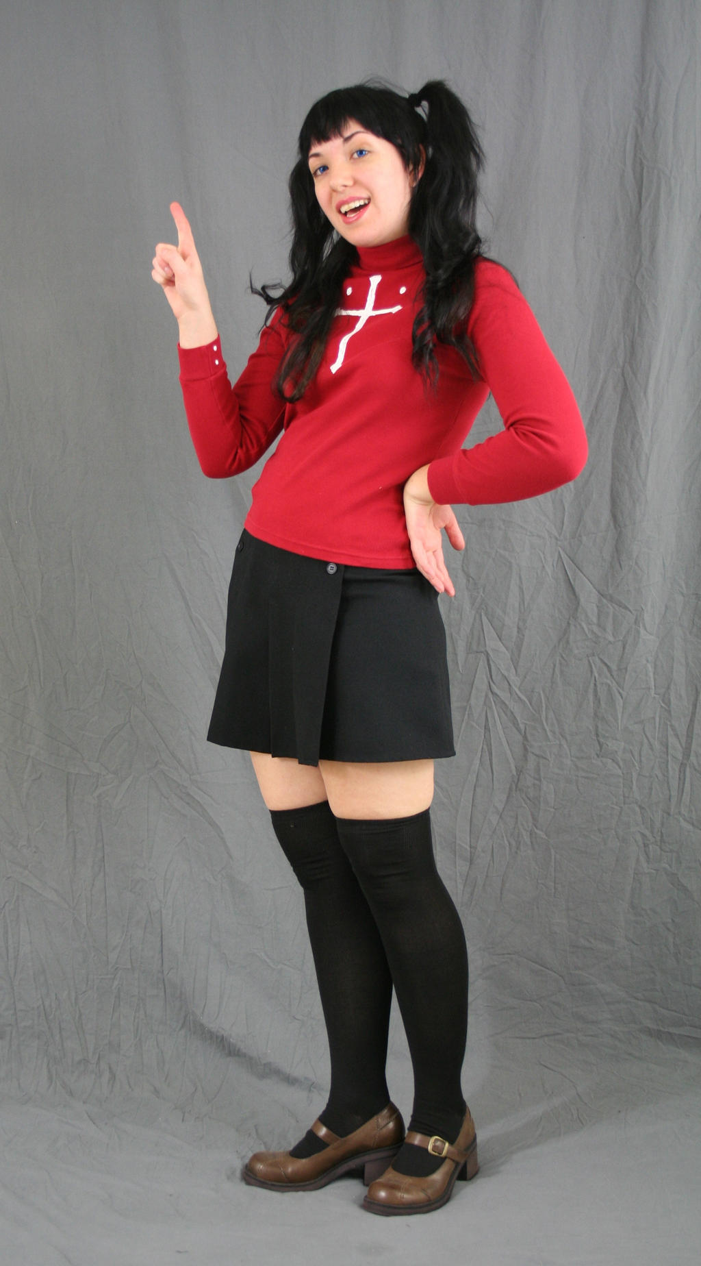 a full shot of a young woman cosplaying a character, possibly from an anime. she's wearing a red longsleeved sweater with a white symbol on the chest, a black pleated skirt, black kneehigh socks, and brown mary jane shoes. her hair is styled in pigtails, and she's pointing upwards with one hand. the background is a plain grey.