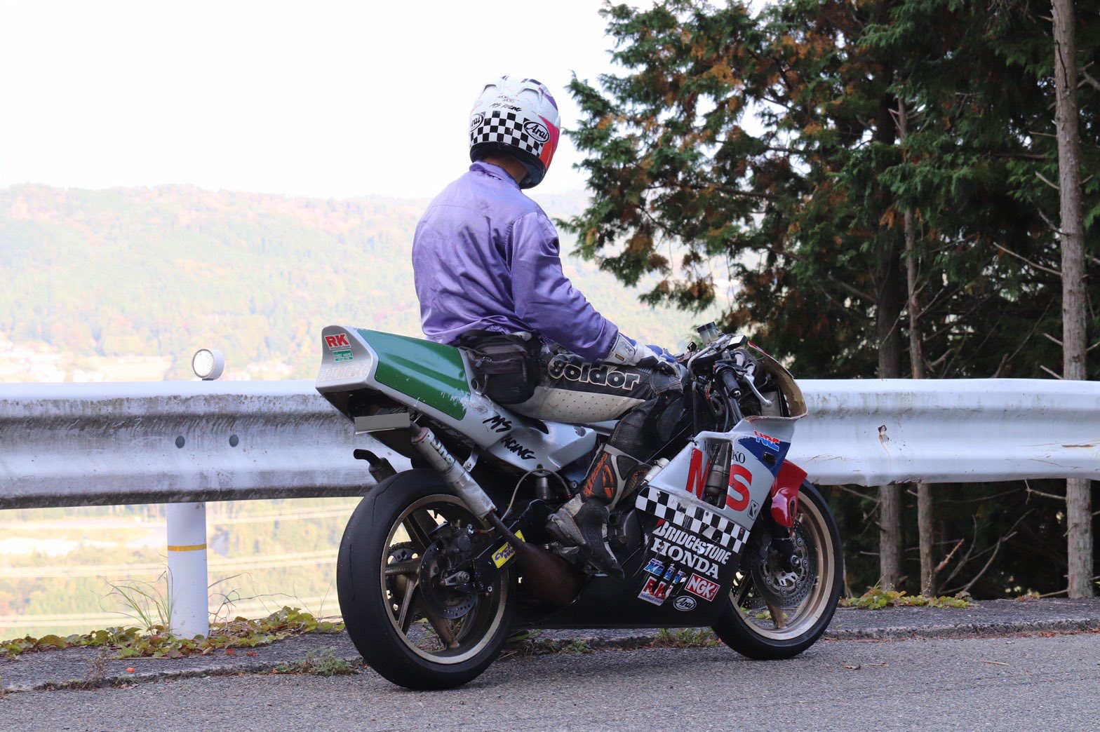 a motorcyclist wearing a helmet is sitting on a motorcycle, overlooking a valley with a mountain range in the background. the road curves around the mountain and there are trees on either side of the road. the motorcycle is a honda and is decorated with racing stickers. the rider is wearing a purple jacket and black pants. the scene evokes a sense of adventure, speed and freedom.