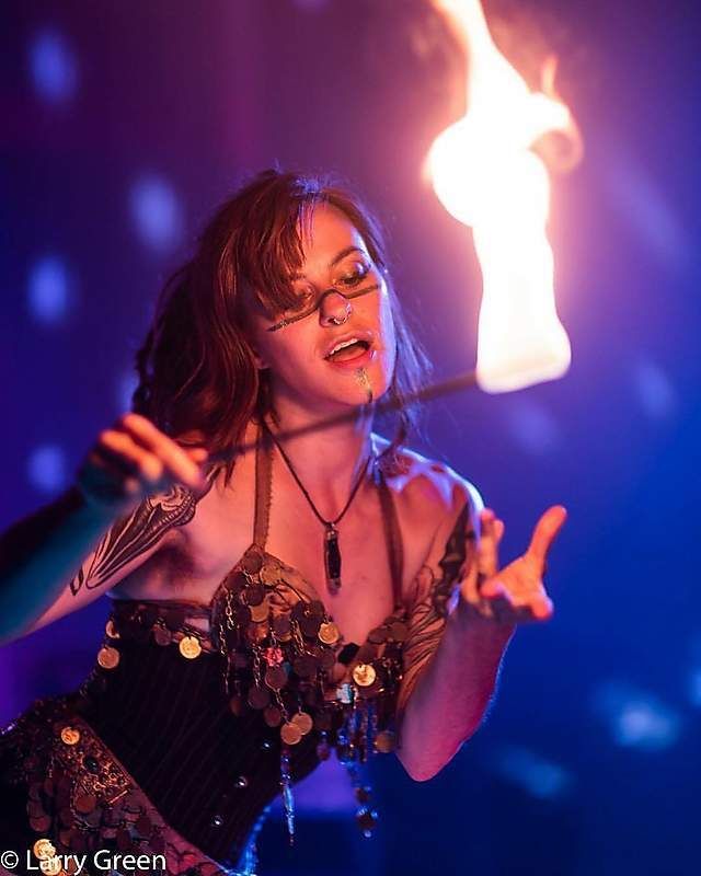 a female performer is dancing on stage and spinning a flaming torch. the stage is dark and lit up with colorful lights. the performer is wearing a beautiful costume and has long hair and makeup.
