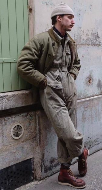 a man in a beanie, jacket, and overalls, leans against a wall in a city. he is wearing boots and looks fashionable. the image is shot from the waist up.