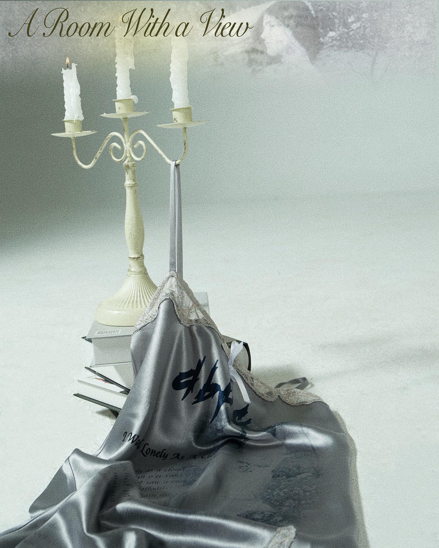 grey silk slip dress with a print lies draped on a surface next to a vintage candle holder with three lit candles.  a stack of books is also visible beneath the candle holder. the background is blurred and features a faded image that seems reminiscent of a painting or illustration; the title "a room with a view" is written across the top.  the overall style of the image is elegant, romantic, and evocative.