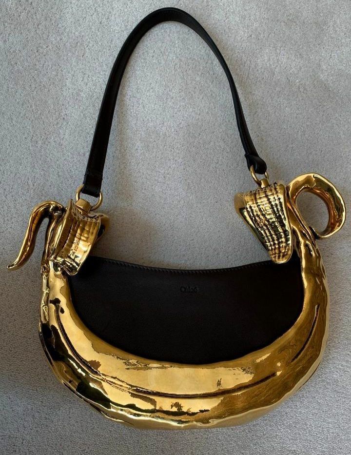 a gold, crescentshaped chloé handbag is shown on a gray surface. it has a black leather strap that attaches to the gold handle via gold loops. each side of the purse has a shell design as the handle.