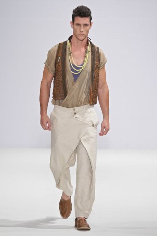 a man is walking towards the camera, dressed in a fashionforward outfit. he is wearing a tan, shortsleeved shirt under a dark brown vest. around his neck, he is wearing a yellow necklace. he is also wearing a pair of light beige, wraparound trousers. on his feet, he is wearing brown shoes. the background is a plain white.