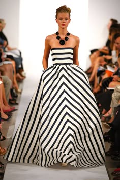 a fashion model walks down a runway in a black and white striped dress. she is wearing a black necklace and has a neutral makeup look.