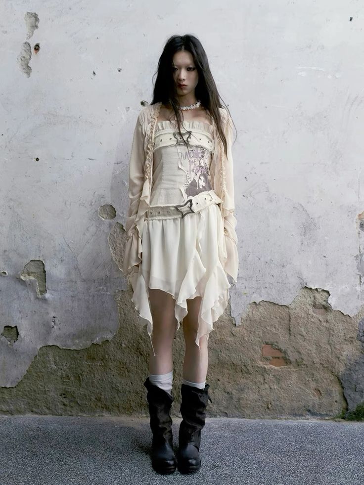a fulllength shot of a young woman with long dark hair modeling an offwhite, asymmetrical dress and a matching cardigan. the dress features ruffles and is worn over a corsetlike top with a graphic print. the model is accessorized with a belt and dark brown, ankleheight boots with a slight foldover detail. she is standing against a heavily textured, distressed, grey wall, which serves as a backdrop to highlight the clothing. the overall style evokes a blend of gothic, alternative, and vintage aesthetics. the mood is moody and somewhat melancholic.