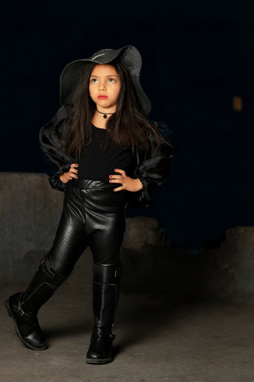 a fullbody shot of a young girl confidently posing in a stylish allblack outfit.  she is wearing a widebrimmed black hat, a black puffedsleeve top, black leather leggings, and tall black boots. the girl's long dark hair is loose, and she strikes a pose with her hands on her hips. the background is dark and slightly out of focus, drawing attention to the girl and her attire. the overall mood is one of edgy fashion and cool confidence.