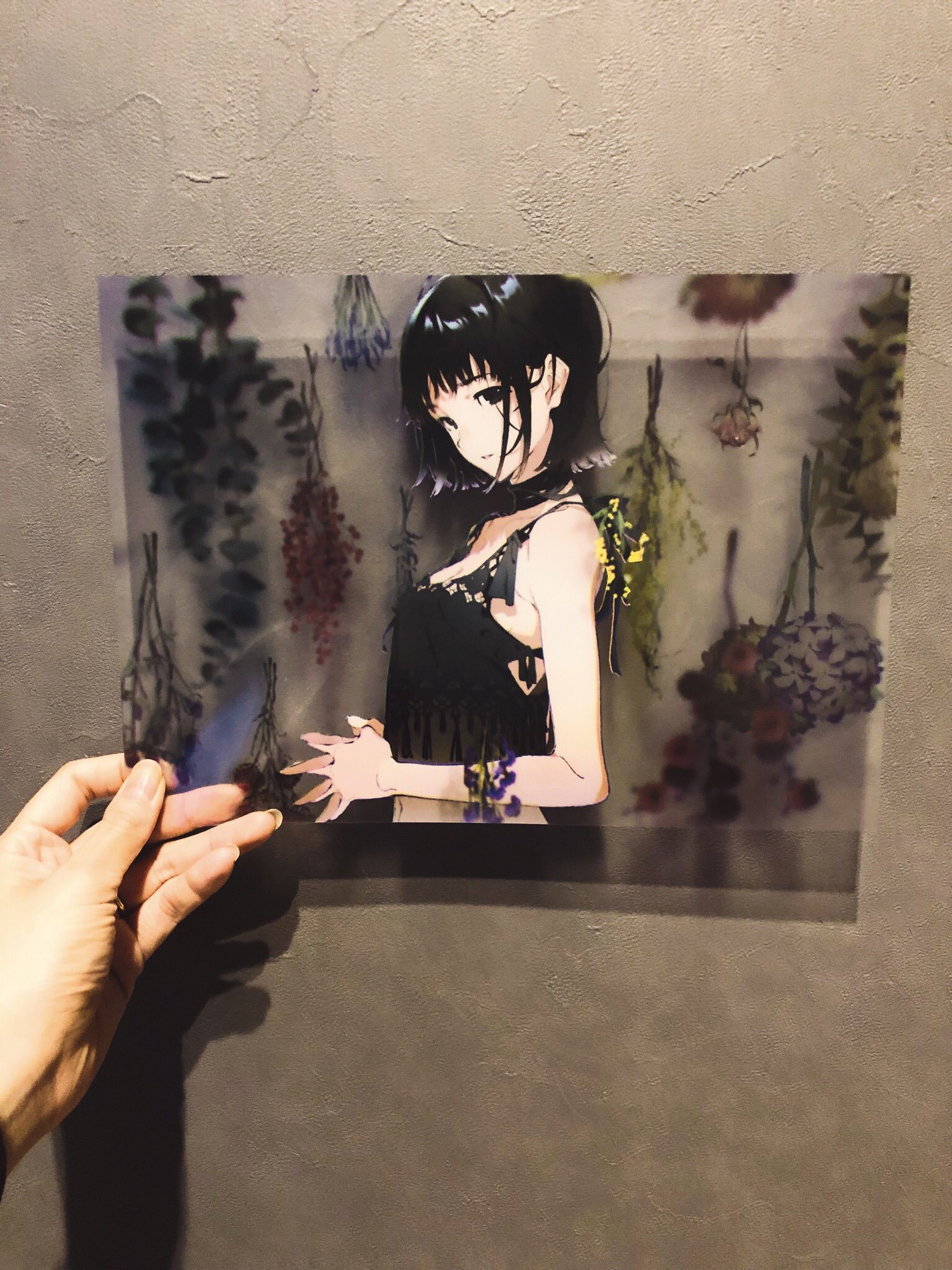 a hand holds up a transparent poster featuring an anime girl with short black hair. she is wearing a black top and standing in front of a wall adorned with hanging flowers.