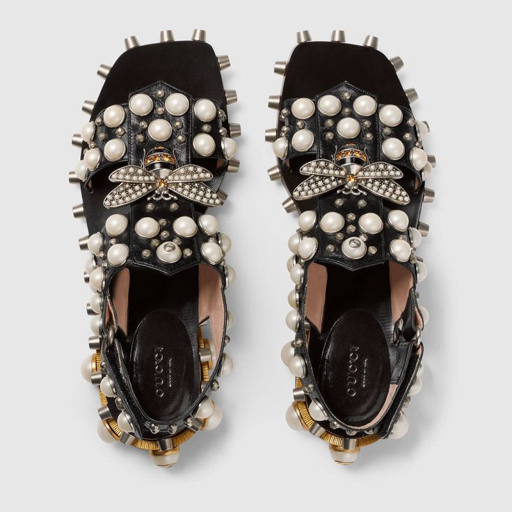 a pair of gucci women's black leather sandals are shown from a bird'seye view. the sandals are adorned with pearls, studs, and a bee detail.  the shoes are positioned on a white background.  the brand label is visible inside each shoe.