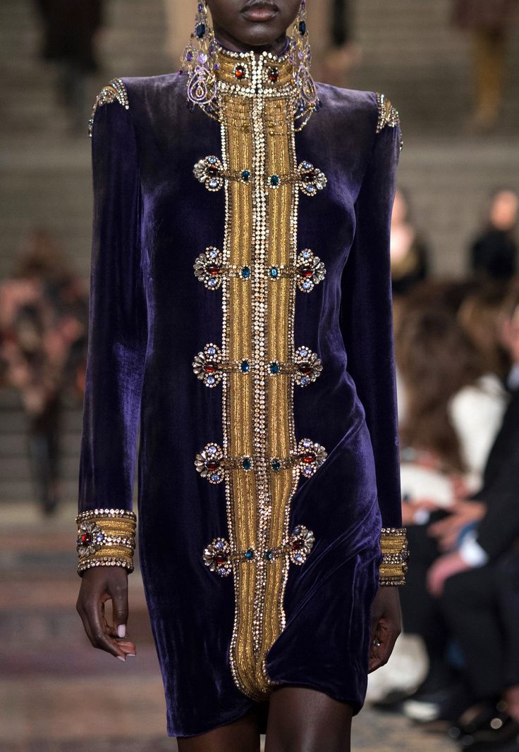 loseup view of a model showcasing a stunning purple velvet dress on a fashion runway. the dress features intricate gold embroidery and embellishments, including various colored jewels, creating a luxurious and regal look. the model's dark skin contrasts beautifully with the rich colors of the attire. the background is blurred, indicating a runway setting with other individuals partially visible. the image focuses on the dress's intricate details and the model's poised stance.